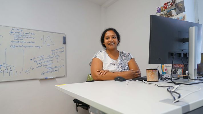 Shuba standing behind a white desk, with her arms relaxing on it in a crossed manner. She is smiling into the camera. On the desk, you can see her computer setup and a big coffee mug. A whiteboard with formulas, graphs and other notes is hanging at the back of the room. The wall next to her desk is decorated with a colorful variety of post cards.